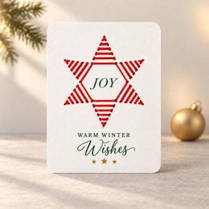 Puede incluir: Una tarjeta de felicitación rectangular blanca con un diseño de estrella roja bordada en el centro y la palabra JOY impresa en verde oscuro. Debajo de la estrella, el texto WARM WINTER WISHES está impreso en verde oscuro, seguido de tres estrellas doradas.