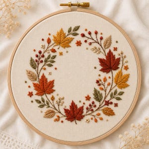 May include: A circular wooden embroidery hoop containing a cream-coloured fabric panel. The fabric features a hand-stitched wreath design composed of vibrant autumn maple leaves in shades of red, orange, and gold, accented with small berries and green foliage.