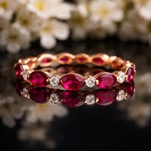 Könnte beinhalten: Ein roségoldener Eternity-Ring mit oval geschliffenen Rubinen und runden Diamanten. Das Design des Rings zeigt abwechselnd Edelsteine, wodurch ein durchgehendes Band aus Farbe und Glanz entsteht. Der Ring wird auf einer reflektierenden Oberfläche präsentiert.