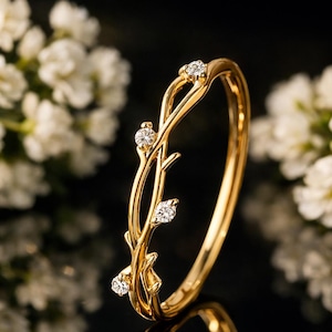 Pode incluir: Um anel de ouro com um delicado design de videira, adornado com pequenos diamantes brilhantes. O anel é apresentado sobre um fundo escuro, com flores brancas ao fundo, criando um contraste que realça os detalhes intrincados.