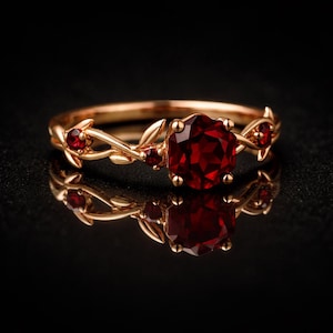 May include: A gold-colored ring with a large, round, deep red gemstone at the center. The ring features smaller red gemstones and a delicate vine and leaf design. The ring is displayed on a reflective black surface.