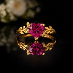 May include: A gold ring with a heart-shaped, magenta gemstone. The ring features a leaf design on the band and around the gemstone. The ring is displayed on a reflective black surface.