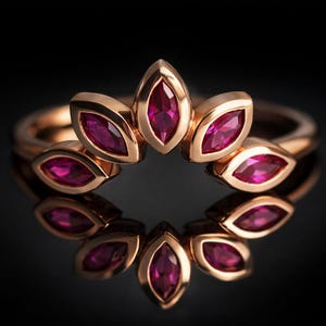 May include: A rose gold ring with a floral design, set with vibrant red marquise-cut gemstones. The ring's design is mirrored in its reflection, creating a symmetrical composition. The ring is displayed against a black background.