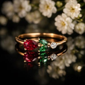 Può includere: Un anello d'oro con una gemma rossa a forma di pera, pietre verdi a forma di foglia e un piccolo diamante. L'anello è posto su uno sfondo scuro con fiori bianchi.