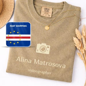 Op de afbeelding: Khaki T-shirt met een camera-afbeelding en de naam "Alina Matrosova, videograaf" erop gedrukt. Een gouden ketting en een strohoed zijn ook zichtbaar. Het shirt is gemaakt van een zacht materiaal.