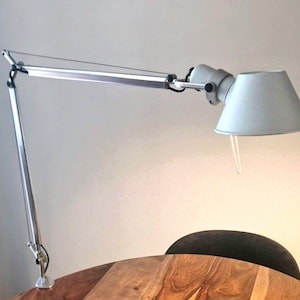 May include: A silver-toned articulated desk lamp with a long, adjustable arm and a conical shade. The lamp features a tension cable system for positioning and a clamp base that secures to a surface. A small white pull cord hangs from the shade.