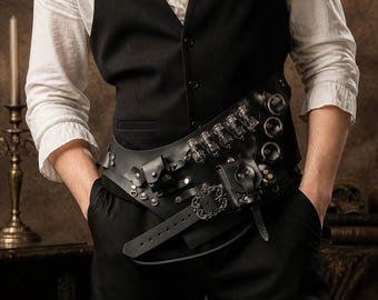 Puede incluir: Un cinturón utilitario de cuero negro sobre una camisa blanca y un chaleco negro. El cinturón presenta varios frascos de vidrio pequeños, dos bolsas de cuero adjuntas, herrajes de metal plateado y una hebilla decorativa con filigrana.