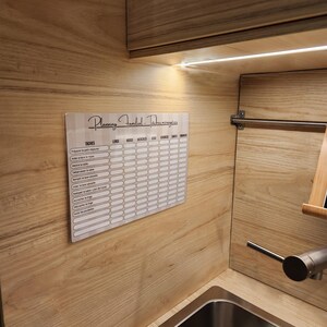 May include: A rectangular white acrylic family chore planner board with black text and a grid layout. The header reads Planning Familial - T&acirc;ches m&eacute;nag&egrave;res, with columns for days of the week and rows for various household tasks in French.