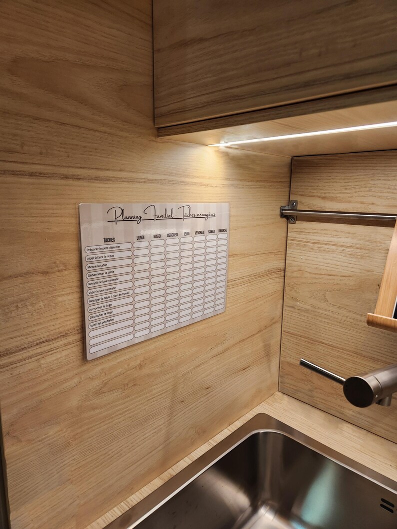 May include: A rectangular white acrylic family chore planner board with black text and a grid layout. The header reads Planning Familial - T&acirc;ches m&eacute;nag&egrave;res, with columns for days of the week and rows for various household tasks in French.