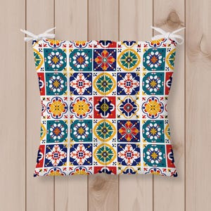 May include: A square seat cushion featuring a repeating tile pattern in vibrant shades of teal, yellow, red, and dark blue on a white background. The cushion includes white fabric ties at the top two corners for securing it to a chair.
