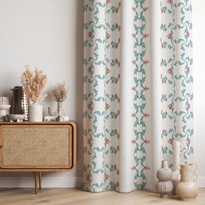 Può includere: Tenda color crema con un motivo floreale ripetuto in verde acqua e rosso. Il design presenta un bordo verticale verde acqua con accenti floreali. La tenda è appesa davanti a un muro bianco, con un mobile in legno e vasi decorativi visibili.