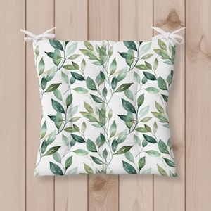 May include: Square cushion with a white background and a repeating pattern of green leaves. The leaves vary in shades of green and are arranged in a natural, flowing design. White ties are attached to the corners.