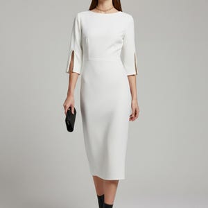 Pode incluir: Vestido midi branco com decote barco e mangas três quartos. As mangas têm detalhes dourados decorativos nos punhos. O vestido é combinado com botins pretos e uma clutch preta. A modelo usa um colar dourado.