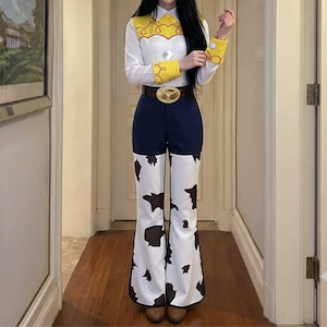 May include: A long-sleeved white shirt with yellow shoulder and cuff accents featuring a red rope pattern, paired with dark blue trousers that transition into white flared pant legs with a black cow-print pattern and a brown belt with a gold buckle.