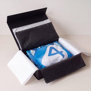 May include: A blue and white jersey featuring a large number 4 graphic on the front, neatly folded inside a black gift box lined with white and black tissue paper. The box has a hinged lid and is open to display the garment inside.