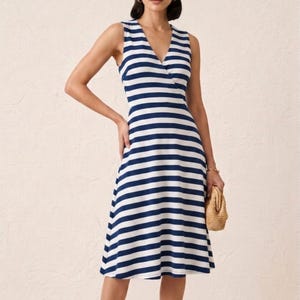 May include: Sleeveless, navy blue and white striped dress with a V-neckline. The dress has a wrap-style bodice and a flared skirt. A small woven handbag is held in the hand.