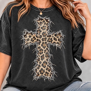 May include: A charcoal gray short-sleeve t-shirt featuring a large, central cross graphic with a brown and black leopard print pattern. The cross design has a distressed, frayed-edge appearance, and the shirt has a faded, washed-out texture.