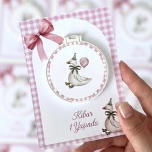 May include: A rectangular card with a pink and white gingham border featuring a circular embroidery hoop frame. The design includes a white goose wearing a party hat and green bow, holding a pink balloon. The text reads Kibar 1 Yaşında.