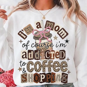 Könnte beinhalten: Weißes Sweatshirt mit dem Text "I'm a mom, of course I'm addicted to coffee & shopping". Die Grafik zeigt einen Kaffeebecher mit rosa Schleife und Leopardenmuster. Das Design hat auch Sterne und einen schwarzen Stern.