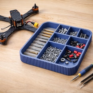 May include: A blue parts organiser filled with screws, bolts, and small components. A black drone frame is in the background. A yellow and blue screwdriver and black tweezers are also present, suggesting a hobbyist or repair theme.