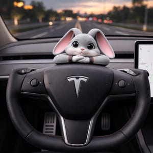 May include: A gray plush bunny car accessory with pink inner ears, resting on a black steering wheel. The bunny has large, expressive eyes and is positioned above the Tesla logo. The steering wheel has red stitching.