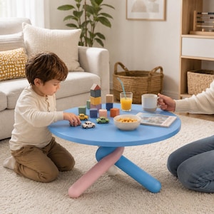 Puede incluir: Una mesa redonda azul claro para niños con un diseño único de tres patas en rosa y azul. La mesa contiene coches de juguete, bloques y un cuenco con aperitivos. Un niño juega con los juguetes. La mesa tiene unos 61 cm de diámetro.