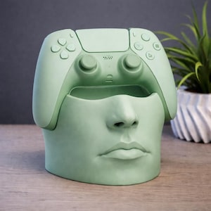 May include: A light green decorative planter shaped like a human face, with a video game controller design on top. The planter is made of a solid material and has a smooth finish. The controller design includes buttons and directional pads.