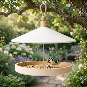 Puede incluir: Un comedero para pájaros moderno, con un techo cónico blanco y una base circular de madera, suspendido de una rama de árbol. Un pequeño pájaro con plumas azules, amarillas y verdes se alimenta de semillas. El comedero está en un jardín.