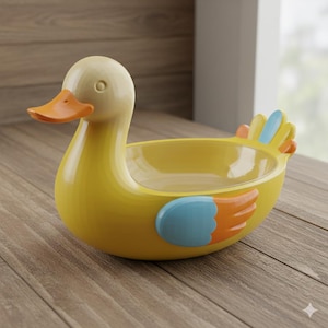 May include: A yellow duck-shaped decorative bowl with an orange beak and blue and orange wing accents. The bowl has a clear interior and is sitting on a wooden surface. The duck has a smooth, glossy finish.