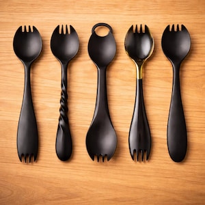 May include: Five black serving spoons with unique designs are arranged on a light wood surface. One spoon has a twisted handle, another has a gold-colored bowl, and one has a ring on the handle. The spoons have a fork-like end.