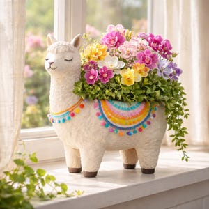 May include: A decorative llama planter, filled with a vibrant array of flowers. The off-white ceramic llama features a smiling face and a blanket with a rainbow design and pom-poms. The flowers include pink, yellow, white, and purple blooms.