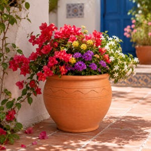 Op de afbeelding: Een grote terracotta bloempot vol levendige bloemen. Het arrangement omvat rode bougainvillea, gele, paarse en witte bloesems. De pot heeft een decoratief golvend lijnenontwerp en staat op een terras met terracotta tegels.