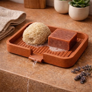 Op de afbeelding: Een terracotta zeepbakje met een ingebouwde afvoer, met een bruin stuk zeep en een natuurlijke luffa spons. Water stroomt uit de afvoer. Het bakje staat op een gespikkelde ondergrond, met een handdoek en kleine planten op de achtergrond.