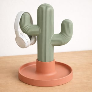 Op de afbeelding: Een groene cactusvormige hoofdtelefoonstandaard met een ronde, terracotta-kleurige basis. De standaard houdt een paar witte hoofdtelefoons vast. De cactus heeft een geribbelde textuur en is ontworpen om hoofdtelefoons vast te houden. De standaard staat op een houten oppervlak.