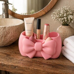 Op de afbeelding: Een roze strikvormige organizer bevat schoonheidsproducten, waaronder een tube getinte moisturizer, concealer en lippenstift. De organizer staat op een houten oppervlak in de buurt van een wastafel en een vaas met bloemen.