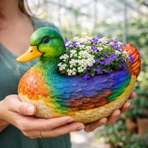 Puede incluir: Una colorida maceta con forma de pato, llena de flores blancas y moradas. El pato tiene un pico amarillo, una cabeza verde y un cuerpo pintado con vibrantes tonos arcoíris. La maceta se sostiene en las manos de una persona, mostrando su diseño decorativo.