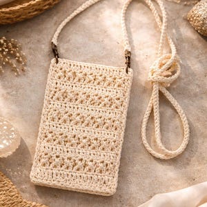 May include: Cream-colored crochet phone bag with a long strap. The bag features a textured, open weave design and metal hardware. The strap is knotted at the end. The bag is shown on a neutral surface with other decorative items.