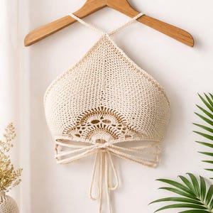 May include: Cream-colored crochet halter top with a decorative openwork design. The top is hanging on a wooden hanger. The halter top has a tie closure at the waist and a crisscross strap design. The top is displayed against a white wall with a palm leaf.
