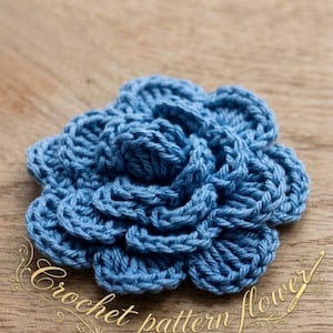 May include: A close-up of a handmade, light blue crochet flower. The flower has multiple layers of petals, creating a textured, three-dimensional effect. The words "Crochet pattern flower" are written in cursive.