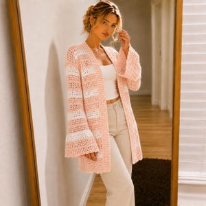 Könnte beinhalten: Eine langärmelige, vorne offene Häkelstrickjacke mit horizontalen Streifen in zartem Pfirsich und Weiß. Das Kleidungsstück hat weite, ausgestellte Glockenärmel und ein strukturiertes Lochstrickmuster, getragen über einem weißen Top und beigen Hosen.