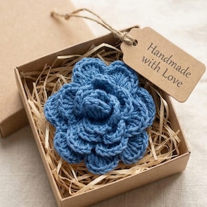 Può includere: Un fiore all'uncinetto blu fatto a mano in una scatola di cartone con imbottitura a forma di paglia. Un'etichetta marrone con le parole "Handmade with Love" è attaccata alla scatola con dello spago. Il fiore ha un design dettagliato e strutturato.