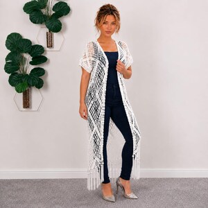 May include: A long, white, open-front crochet cardigan featuring a diamond-patterned knit design, short sleeves, and a fringed hemline. The garment is worn over a dark blue tank top and dark blue fitted trousers, creating a layered appearance.