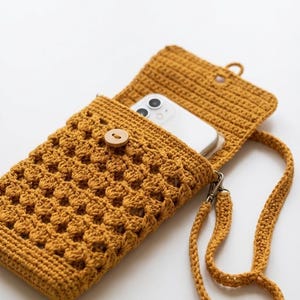 Puede incluir: Bolso de teléfono de ganchillo amarillo mostaza con cierre de botón de madera y correa a juego. El bolso tiene un patrón de tejido abierto y texturizado y un cierre plateado. Un teléfono inteligente blanco es visible dentro de la bolsa abierta.
