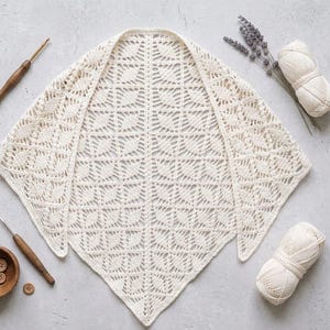 May include: A cream-coloured, open-weave crochet shawl with a geometric leaf pattern. The shawl is laid flat, with a wooden crochet hook, yarn, and wooden buttons nearby. A sprig of lavender and a ball of yarn are also present.