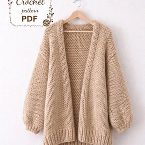Può includere: Un cardigan beige con davanti aperto e maniche lunghe e voluminose è appeso a una gruccia di legno. Il maglione a maglia grossa ha un orlo e polsini a coste. L'immagine include il testo "Crochet pattern PDF" in una cornice decorativa.