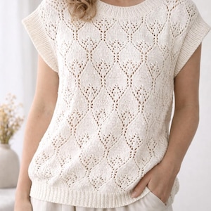 May include: Cream-colored, short-sleeved knit top with a detailed diamond pattern. The top has a crew neck and ribbed hem. The model is wearing the top with light-colored pants. The top is a stylish addition to any wardrobe.