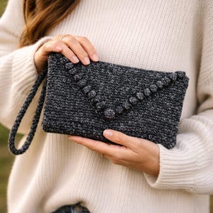 Könnte beinhalten: Dunkelgraue gehäkelte Clutch mit Handschlaufe. Die Clutch hat eine dekorative Reihe von Bommeln entlang der Klappe. Die handgefertigte Tasche wird vor einem cremefarbenen Pullover und Jeans gehalten.