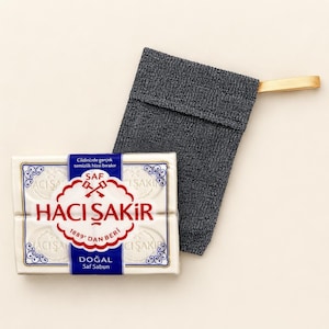 Puede incluir: Una pastilla de jabón blanca rectangular Hacı Şakir con texto de marca en azul y rojo y un gráfico de dos llaves cruzadas, junto a una bolsa exfoliante texturizada de color gris oscuro con una trabilla de color beige claro.