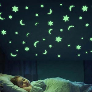 Peut inclure: Une chambre sombre avec un enfant endormi sous un plafond décoré d'autocollants en forme d'étoiles et de croissants de lune verts brillants. Les autocollants de différentes tailles créent une lumière ambiante douce.