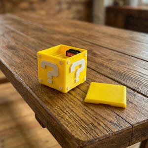 Peut inclure: Une boîte de rangement jaune imprimée en 3D avec des points d'interrogation blancs sur les côtés, ressemblant à un objet de jeu vidéo. La boîte est ouverte, révélant une cartouche de jeu à l'intérieur, et possède un couvercle jaune séparé.